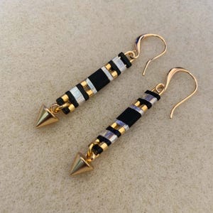 May include: A pair of gold-toned earrings with a black and gold beaded design. The earrings have a long, rectangular shape with a gold cone-shaped pendant at the bottom. The ear hooks are gold-toned.