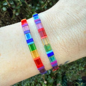 Puede incluir: Dos pulseras elásticas compuestas por cuentas de vidrio cuadradas, translúcidas y multicolores. Las cuentas presentan un espectro vibrante de tonos rojos, naranjas, amarillos, verdes, azules, morados e irisados en un patrón lineal.