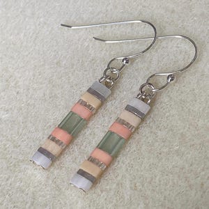 Op de afbeelding: Een paar oorhangers met zilveren haken. De oorbellen zijn gemaakt van kleine, kleurrijke kralen in roze, groen, beige, grijs en transparant. De kralen zijn in een lineair patroon gerangschikt.