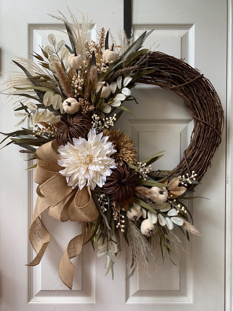 May include: A fall-themed wreath features a brown vine base adorned with creamy white and beige artificial flowers, including dahlias and dried seed pods.  Subtle accents of dried grasses and wheat add texture. A burlap bow complements the neutral color palette. The wreath is approximately 24 inches in diameter and ideal for autumnal decor.