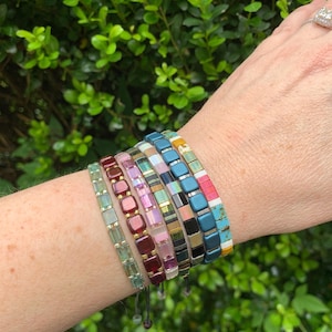 May include: A collection of colorful tile bracelets in various shades of blue, pink, green, and gold. The bracelets are made of small, square tiles strung together on a cord. The bracelets are displayed on a wrist against a backdrop of green foliage.