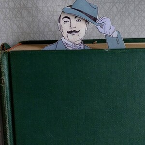 May include: A green book with a man's face peeking out from behind the top edge. The man is wearing a blue hat and a bow tie. He is smiling and looking at the camera.