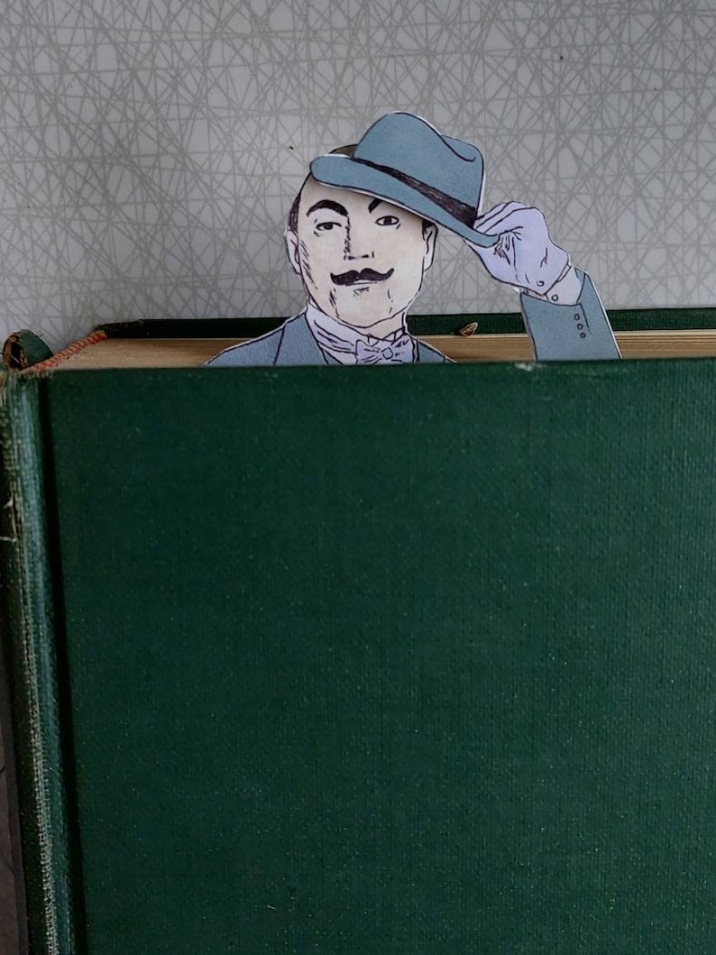 May include: A green book with a man's face peeking out from behind the top edge. The man is wearing a blue hat and a bow tie. He is smiling and looking at the camera.