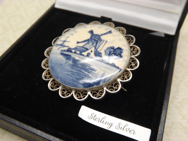 Pode incluir: Um broche de filigrana de prata com um camafeu de porcelana azul e branco que representa um moinho de vento. O camafeu est&aacute; engastado em uma moldura de filigrana de prata e o broche &eacute; marcado com "Sterling Silver".