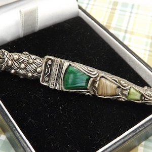 May include: A silver-toned metal brooch shaped like a traditional Scottish dirk dagger. The handle features a textured pattern with a round amber-colored stone at the top. The blade section is inlaid with green malachite, tan agate, and light green stone.