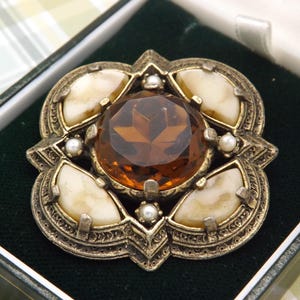 May include: A square-shaped brooch with an antiqued gold-tone metal frame featuring a large, faceted amber-coloured central stone, four cream-coloured curved cabochons at the corners, and four small white pearls set between the cabochons.