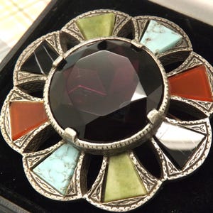 Op de afbeelding: Een vintage broche met een grote, facetgeslepen, donkerpaarse middensteen. De broche heeft een zilverkleurig metalen frame met kleurrijke driehoekige inzetstukken van rode, groene, zwarte en turkooizen stenen. De broche staat tegen een zwarte achtergrond.