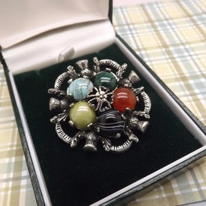Pode incluir: Broche prateado com seis pedras redondas coloridas, em tons de azul, verde, amarelo, vermelho e preto. O broche é definido em um design decorativo floral e apresentado em uma caixa forrada de veludo preto.
