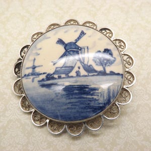 Pode incluir: Um broche de filigrana de prata com um camafeu de porcelana azul e branco que representa uma cena de moinho de vento holand&ecirc;s.
