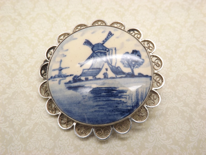 Pode incluir: Um broche de filigrana de prata com um camafeu de porcelana azul e branco que representa uma cena de moinho de vento holand&ecirc;s.