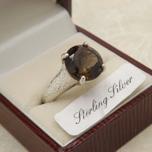 Peut inclure: Une bague en argent avec une grande pierre précieuse ronde de couleur brune. La bague est dans un écrin en velours rouge avec une étiquette blanche qui indique "Sterling Silver".