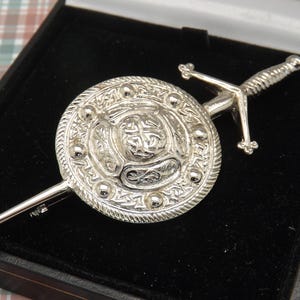 May include: A silver-toned kilt pin featuring a round shield design with intricate detailing and a decorative sword. The pin is presented in a black box, showcasing its ornate craftsmanship. The shield has a diameter of approximately 1.5 inches.