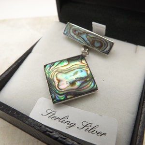 May include: A sterling silver brooch featuring a square abalone shell design. The brooch is presented in a black box with a white label that reads "Sterling Silver".