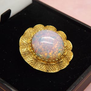 May include: A gold brooch featuring a large, iridescent opal-like stone. The stone is set within a detailed, floral-shaped frame. The brooch is displayed in a black jewellery box, possibly indicating a vintage or antique item.