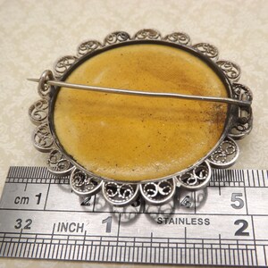 Pode incluir: Um broche de filigrana de prata com um camafeu redondo amarelo. O broche tem 2,5 cm de di&acirc;metro e um fecho de pino.