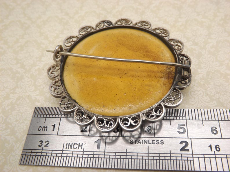 Pode incluir: Um broche de filigrana de prata com um camafeu redondo amarelo. O broche tem 2,5 cm de di&acirc;metro e um fecho de pino.