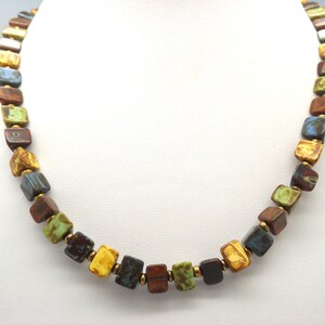 May include: A necklace with square beads in shades of brown, yellow, green, and blue. The beads are linked with gold-coloured connectors. This colourful necklace is displayed against a white backdrop. The necklace is a unique accessory.