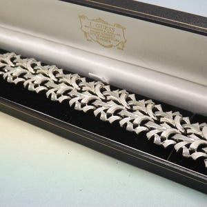 Può includere: Bracciale argentato con un motivo a foglie ripetute, esposto in un cofanetto nero con fodera in raso bianco. Il bracciale è composto da più sezioni collegate, ciascuna con un motivo a foglie stilizzate. La scatola ha un'emblema dorato e testo.