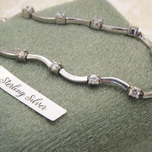 Op de afbeelding: Een sterling zilveren armband met een golvend ontwerp, met meerdere heldere, rond geslepen edelstenen. De armband wordt gepresenteerd op een groene ondergrond, met een label met "Sterling Silver."