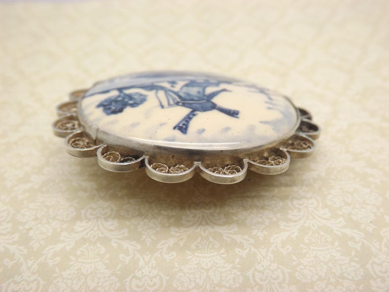 Pode incluir: Um broche de filigrana de prata com um camafeu de porcelana branca com um design de moinho de vento azul e branco.