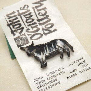 Könnte beinhalten: Eine schwarze und weiße Keramikskulptur einer Highland-Kuh, mit dem Text "John O'Groats Pottery" und den Kontaktdaten des Unternehmens.