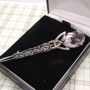 May include: A silver-toned kilt pin with a Celtic knot design and a large, oval, light pink gemstone. The pin is displayed in a black presentation box with a velvet lining, set against a plaid background.
