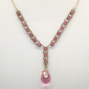 May include: A vintage gold-toned necklace with pink gemstones. The necklace features a delicate chain design with a series of round pink stones and a large, teardrop-shaped pink pendant. The necklace is displayed against a white background.