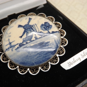 Pode incluir: Um broche de filigrana de prata com um camafeu de porcelana azul e branco que representa um moinho de vento holand&ecirc;s. O camafeu est&aacute; engastado em uma moldura de filigrana de prata. A parte de tr&aacute;s do broche tem as palavras "Sterling Silver" gravadas.