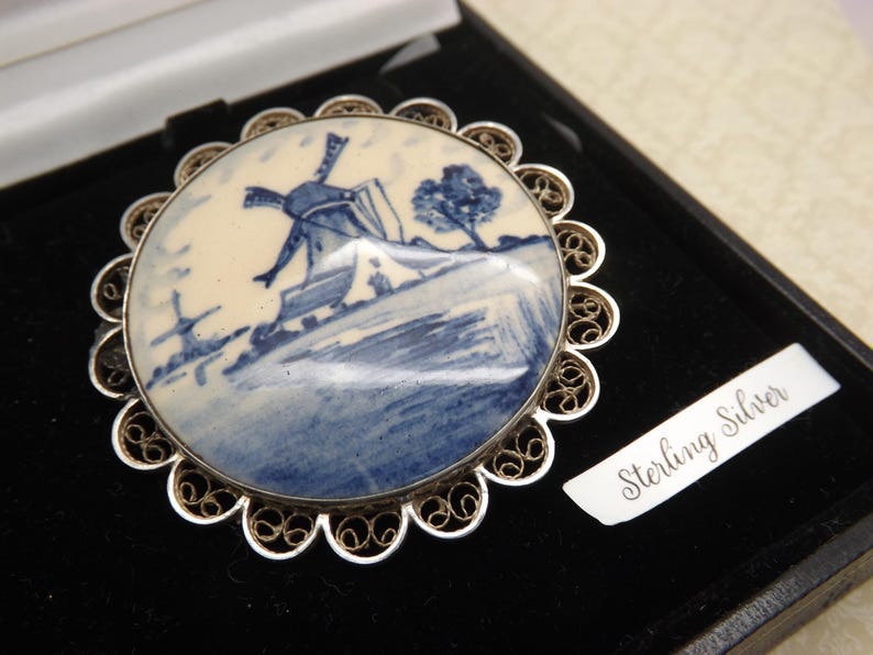 Pode incluir: Um broche de filigrana de prata com um camafeu de porcelana azul e branco que representa um moinho de vento holand&ecirc;s. O camafeu est&aacute; engastado em uma moldura de filigrana de prata. A parte de tr&aacute;s do broche tem as palavras "Sterling Silver" gravadas.