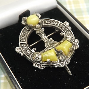 May include: A silver-toned brooch with a circular design, featuring yellow-toned gemstones and intricate detailing. The brooch is presented in a black box, showcasing its ornate craftsmanship and vintage style.