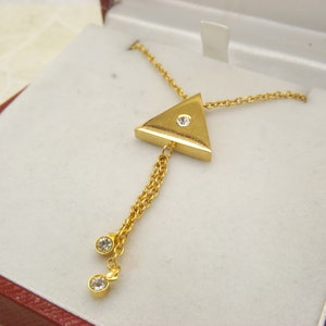 Könnte beinhalten: Eine goldene Halskette mit einem dreieckigen Anhänger, der mit einem einzigen klaren Stein besetzt ist. Der Anhänger hängt an einer zarten Kette mit einem kleinen Kettenquaste mit zwei klaren Steinen am Ende.