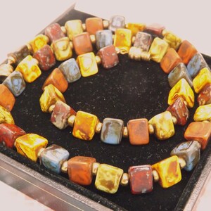 May include: A colourful necklace featuring square beads in shades of yellow, orange, red, and blue. The beads are strung together with gold-coloured spacers. The necklace is displayed on a black velvet surface.