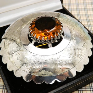 Peut inclure: Une broche circulaire en argent avec un bord festonné et des motifs de chardon gravés. Le centre de la broche est orné d'une grande pierre facettée de couleur ambre, maintenue par un sertissage décoratif en argent à bord festonné.