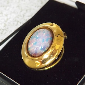 Op de afbeelding: Een goudkleurige ovale broche met een iriserende, opalescente middensteen. De broche is gezet in een gepolijste gouden lijst en rust op een zwart fluwelen oppervlak in een doos. De steen toont tinten blauw, roze en wit.