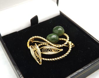 May include: A gold-toned brooch with a decorative design featuring two green, round gemstones. The brooch has a twisted rope-like border and filigree details, presented in a black jewelry box.