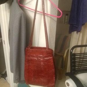 May include: A red handbag with a textured, snake-like pattern hangs from a pink hanger. The bag has a rectangular shape with a long strap. The background includes a gray coat and a glimpse of a shelf.