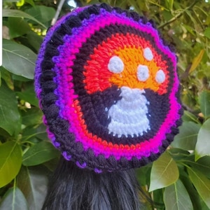 May include: A colorful crochet mushroom hat with a black base, vibrant pink, orange, and purple accents. The mushroom design features a white stem and white spots on the orange cap. The hat is handmade.