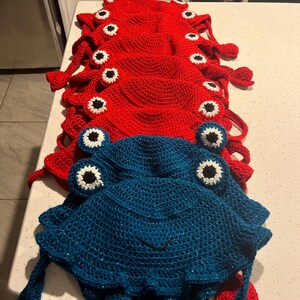 May include: A stack of red crocheted frog hats with black and white eyes, and a blue crocheted frog hat with black and white eyes.