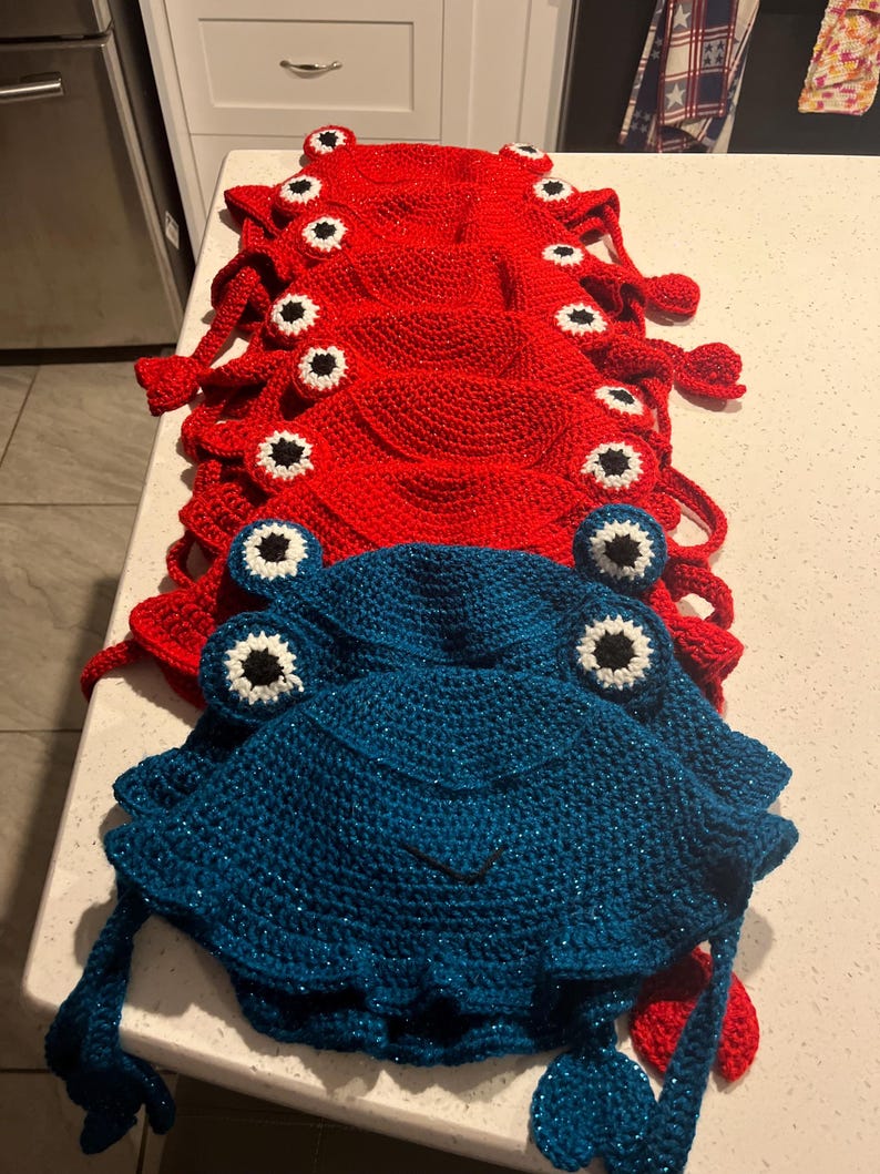 May include: A stack of red crocheted frog hats with black and white eyes, and a blue crocheted frog hat with black and white eyes.