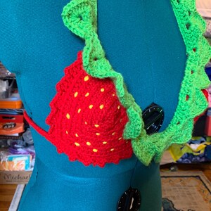 May include: A handmade crochet bikini top in the shape of a strawberry. The top is red with yellow seed details and green leaf trim. The bikini top is displayed on a teal mannequin.