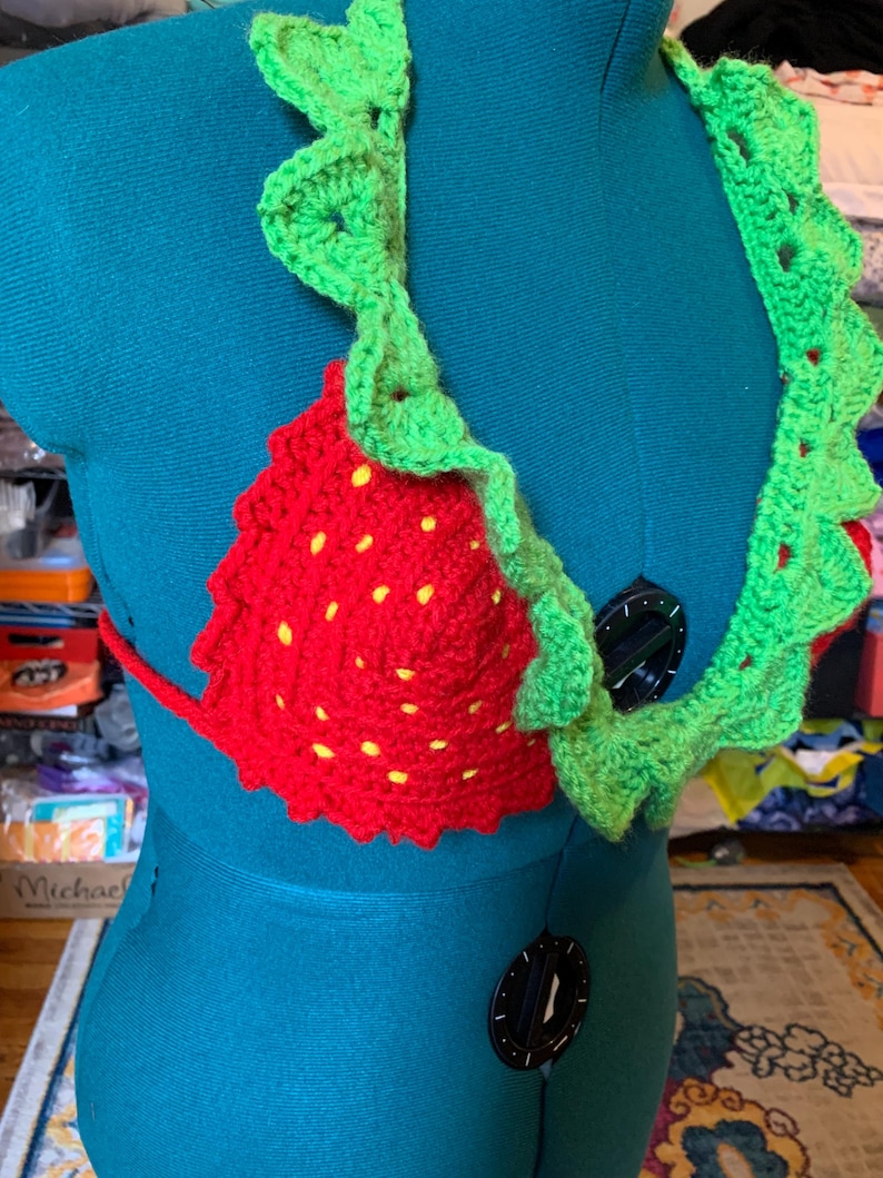 May include: A handmade crochet bikini top in the shape of a strawberry. The top is red with yellow seed details and green leaf trim. The bikini top is displayed on a teal mannequin.