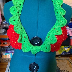 May include: A handmade crochet halter top in a strawberry design. The top features a red bodice with yellow seed details and a green scalloped trim. The halter top is displayed on a teal dress form.