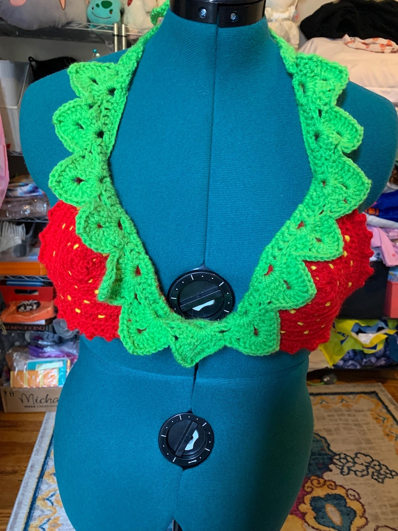 May include: A handmade crochet halter top in a strawberry design. The top features a red bodice with yellow seed details and a green scalloped trim. The halter top is displayed on a teal dress form.