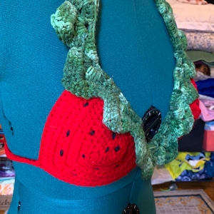 May include: A handmade crochet bikini top in a strawberry design. The top features a red crocheted cup with black seed details and a green leafy trim. The halter straps and neck trim are also green.