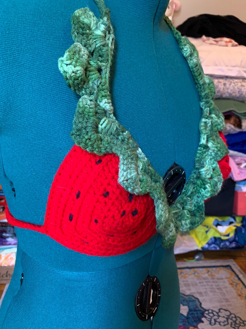 May include: A handmade crochet bikini top in a strawberry design. The top features a red crocheted cup with black seed details and a green leafy trim. The halter straps and neck trim are also green.