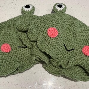 May include: Two handmade green crochet frog hats. Each hat features pink cheek circles, black stitched eyes, and white and black eye accents. The hats have a playful, whimsical design.