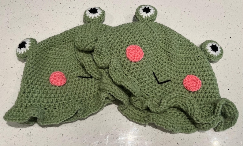 May include: Two handmade green crochet frog hats. Each hat features pink cheek circles, black stitched eyes, and white and black eye accents. The hats have a playful, whimsical design.