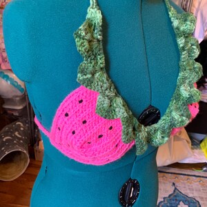 May include: A handmade crochet top in the shape of a watermelon. The top is bright pink with black seed details and green leafy trim. The halter straps and trim are green, creating a fun, summery look.