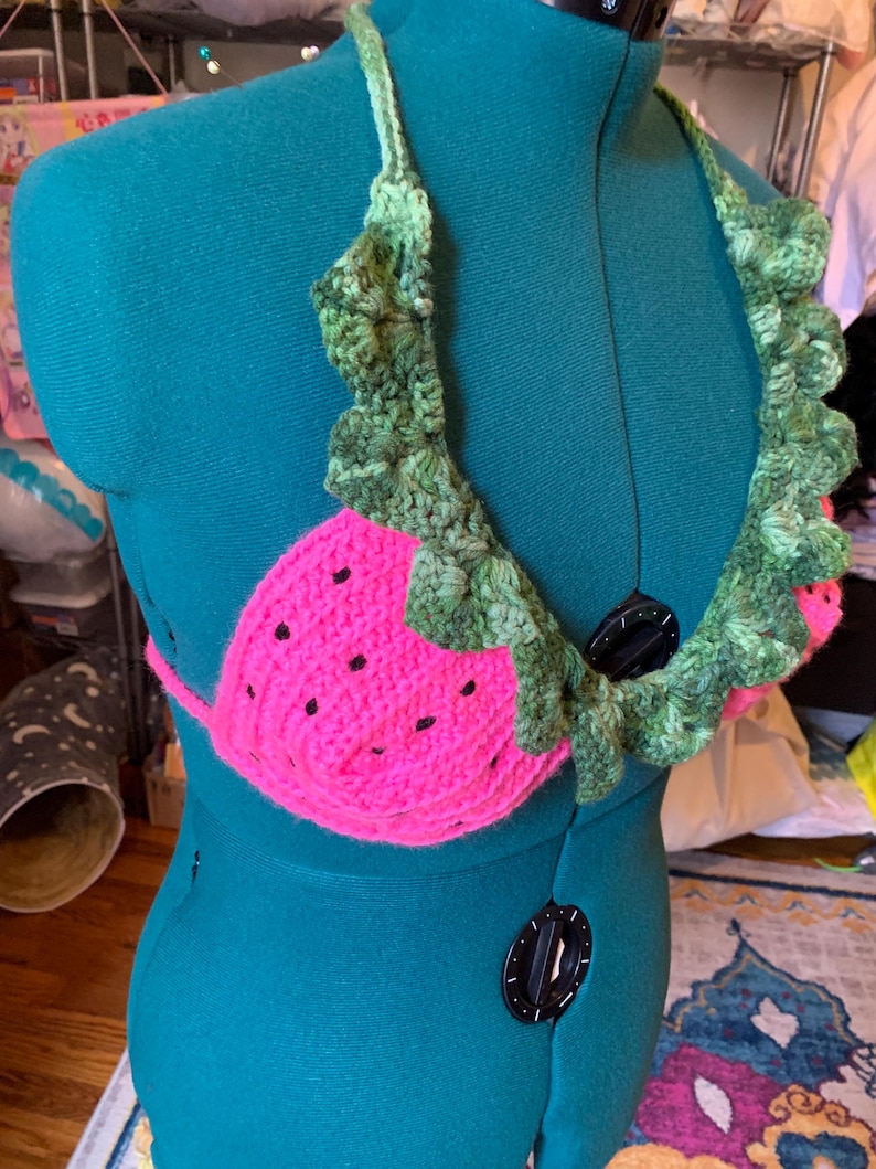 May include: A handmade crochet top in the shape of a watermelon. The top is bright pink with black seed details and green leafy trim. The halter straps and trim are green, creating a fun, summery look.