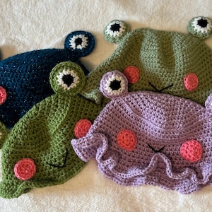 May include: Four handmade crochet frog hats in shades of blue, green, and purple. Each hat features large, round eyes with white and black details, pink cheek circles, and a stitched mouth. The hats are designed to be worn.